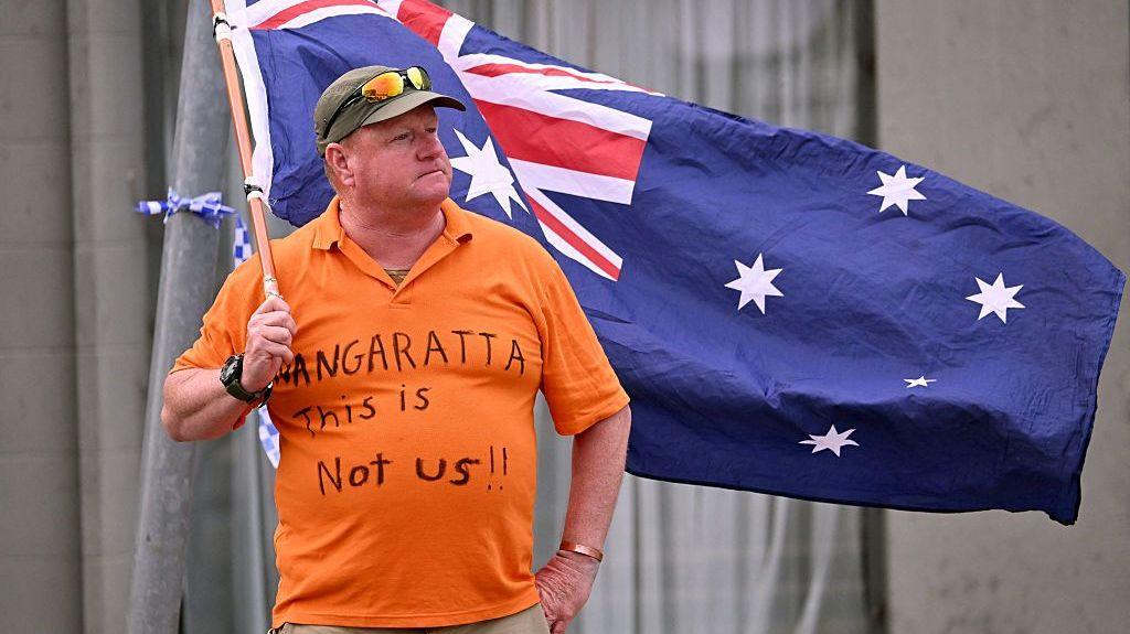 A man wearing an orange t‑shirt reading ‘Wangaratta this is not us!!’ waves an Australian flag