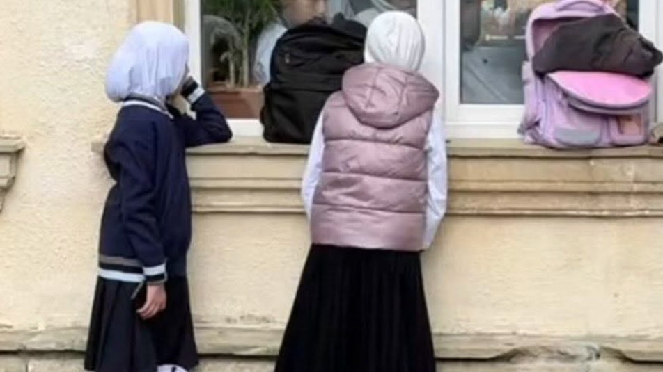 Two hijab-wearing schoolgirls are peering through a school window, with a pink schoolbag placed on the window ledge next to them